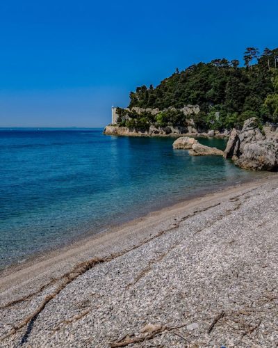 Tranquil rocky beach scene with clear blue waters and lush greenery under a bright sky.